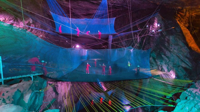 Grŵp o bobl mewn offer diogelwch yn bownsio ar rwydi mewn ogof o dan y ddaear.