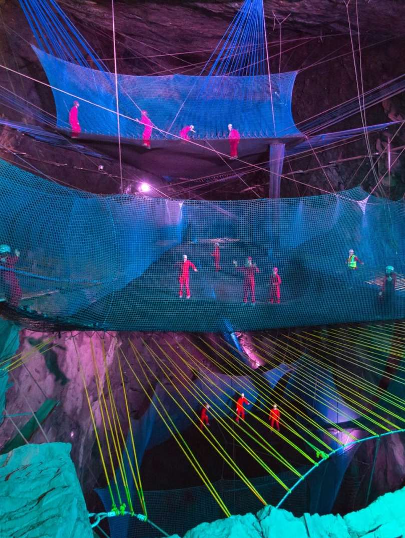 Grŵp o bobl mewn offer diogelwch yn bownsio ar rwydi mewn ogof o dan y ddaear.