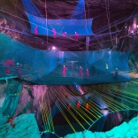 Grŵp o bobl mewn offer diogelwch yn bownsio ar rwydi mewn ogof o dan y ddaear.