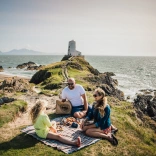 Teulu yn cael picnic ar fryn arfordirol ger goleudy.